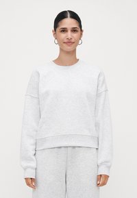 Jovem mulher com cabelo escuro apanhado em um coque, usando brincos de argola prateados, um sweatshirt cinza claro e calças de moletom combinando, de pé contra um fundo branco.