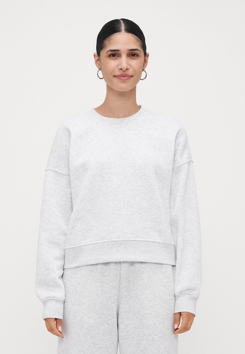 Jovem mulher com cabelo escuro apanhado em um coque, usando brincos de argola prateados, um sweatshirt cinza claro e calças de moletom combinando, de pé contra um fundo branco.