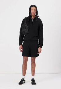 Zwarte pullover met een halflange ritskraag, gecombineerd met zwarte shorts. Model draagt een zwarte schoudertas en zwarte sneakers met witte accenten.