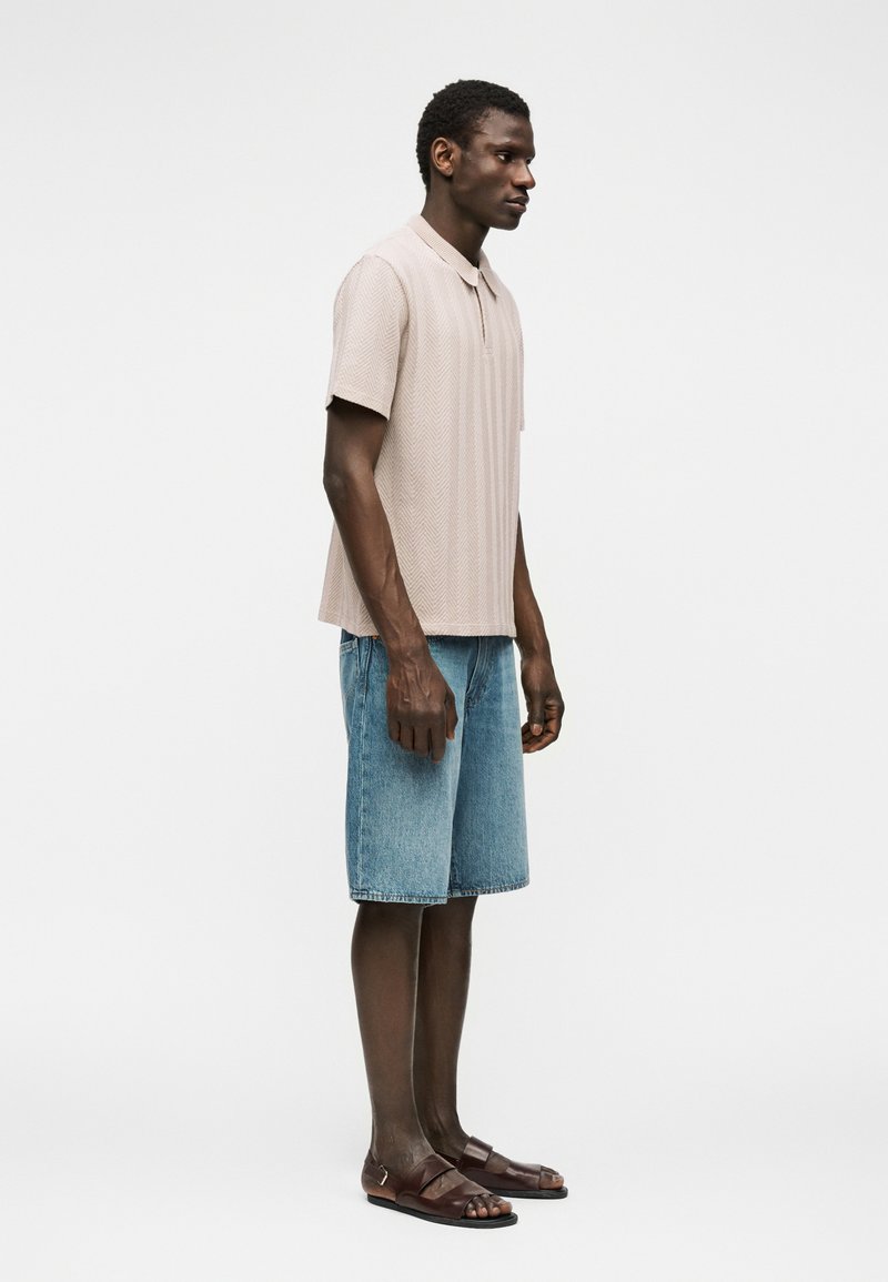 Homme debout en polo beige à manches courtes, short en jean bleu jusqu'aux genoux et sandales marron foncé sur un fond blanc uni.