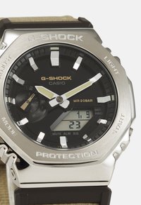 Montre G-SHOCK avec un boîtier en acier inoxydable, un cadran noir, des accents argentés et dorés, et un affichage numérique ; elle est dotée d'un bracelet en résine marron durable.