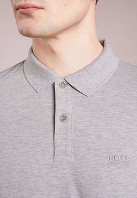 Polo gris à col boutonné, avec un petit bouton vert et un logo "BOSS" brodé sur la poitrine. Texture douce.