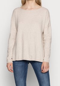 Pull en tricot texturé beige clair, avec un large col et des épaules tombantes, doté de manches longues et d'une coupe décontractée. Associé à un jean bleu.
