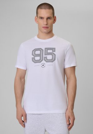 Mężczyzna z krótkimi włosami, ubrany w białą koszulkę z czarną grafiką "95" oraz jasnoszare spodnie, stojący na jednolitym szarym tle.