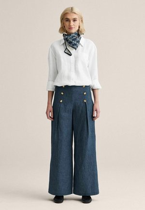 Kvinde iført hvid skjorte med knapper, marineblå bukser med vide ben og guldknapper, sorte sko og mørkt mønstret halstørklæde, stående foran beige baggrund.