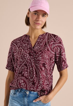 Jeune femme portant une casquette rose, un chemisier bordeaux à manches courtes avec un motif paisley, et un jean bleu, posant avec une main dans la poche.