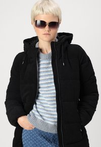 Doudoune en velours côtelé noir avec une capuche, portée sur un pull rayé gris et bleu, associée à un pantalon bleu à motifs, accessoirisée avec des lunettes de soleil.