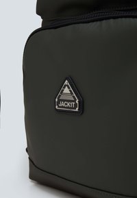 Zielony plecak ma gładką, matową teksturę, trójkątną naszywkę z logo "JACK1T" oraz zamek błyskawiczny wzdłuż górnej krawędzi.