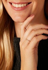 Gouden ring met vijf heldere stenen, met een gebogen ontwerp. Hand rustend nabij een gezicht met schouderlang blond haar. Zwart textiel kledingstuk.