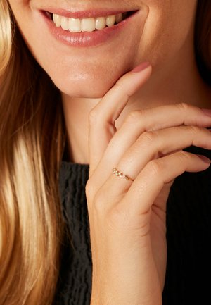 Bague en or avec cinq pierres claires, présentant un design courbé. Main reposant près d'un visage avec des cheveux blonds mi-longs. Vêtement noir texturé.