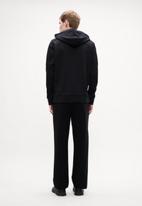 Μαύρη μπλούζα με κουκούλα, υφασμάτινη υφή και ριγέ μανσέτες, σε συνδυασμό με χαλαρούς μαύρους παντελόνες και μαύρα υποδήματα, σε προοπτική από πίσω.