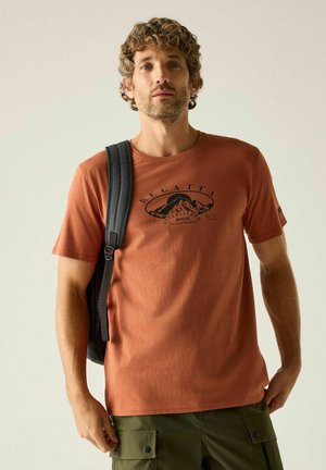 Mann mit lockigem Haar trägt ein rostorangefarbenes T-Shirt mit Berggrafik, grüne Cargoshorts und einen schwarzen Rucksack über einer Schulter.