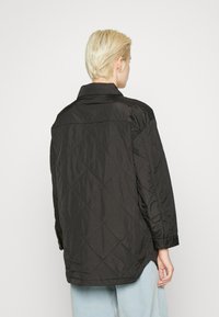 Svart quiltet jakke med en lett, glansfull tekstur. Har krage og lange ermer, med en litt buet kant.