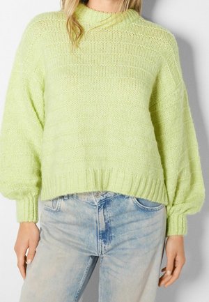 Femme portant un pull en grosse maille vert clair avec des manches bouffantes et un jean taille haute bleu clair.