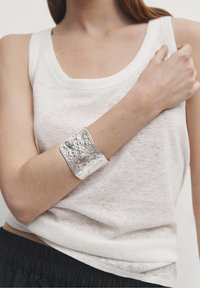 Braccialetto in argento con finitura martellata e texture, indossato su un braccio scoperto, in contrasto con una canotta bianca e una cintura elastica scura.