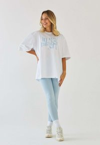 Biała oversize koszulka z niebieskim napisem, jasnoniebieskie legginsy, białe skarpetki i beżowe sneakersy; gładki materiał i luźny krój.