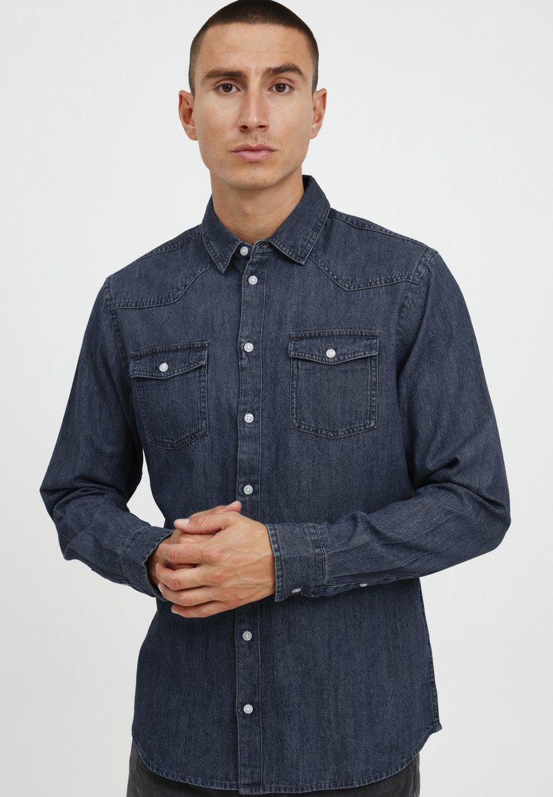 Camisa de denim oscuro con botón en la parte frontal, dos bolsillos en el pecho, mangas largas y un cuello clásico. Presenta costuras en tono y botones plateados.