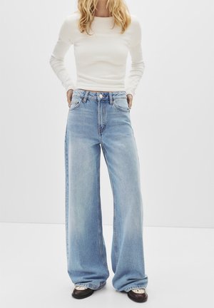 Kvinde iført lyseblå jeans med brede ben og en tætsiddende hvid bluse med lange ærmer, stående med hænderne i lommerne mod en ensfarvet baggrund.