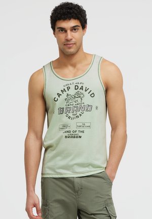 Man with curly hair wearing light green sleeveless tank top with printed text and olive green cargo shorts, standing with hand in pocket.