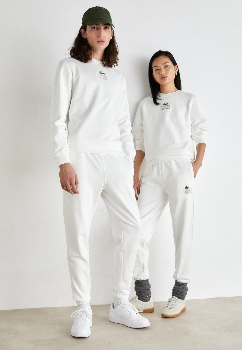 Felpe e pantaloni jogger in cotone bianco con polsini a coste, decorati con loghi ricamati verdi, abbinati a sneakers bianche e beige.