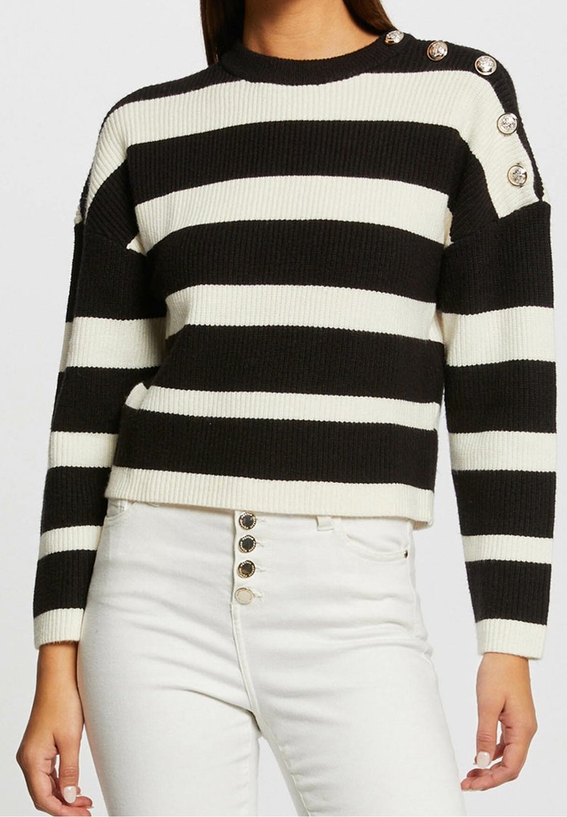 Pull à manches longues rayé noir et blanc avec des boutons argentés sur l'épaule, associé à un pantalon blanc taille haute avec des boutons à l'avant.