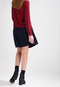 Donna con una blusa lunga bordeaux a maniche lunghe, gonna blu navy, stivali neri alla caviglia e calzini neri, in piedi con la schiena rivolta e i capelli sciolti.