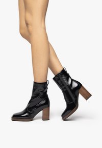 Bottines en cuir verni noir avec un design froncé, talon carré brun et fermeture éclair sur le côté. Texture lisse et finition brillante.
