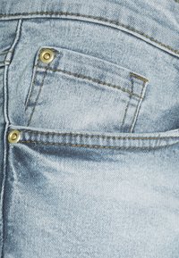 Lysblå denimjeans har en baglomme med gyldne nitter, synlig syning og en lidt slidt tekstur.