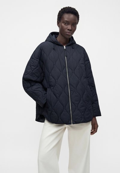 Femme portant une veste matelassée noire à capuche avec fermeture éclair devant et un pantalon blanc à jambes larges, debout devant un fond uni.