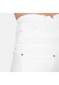 Jean en denim blanc avec un design ajusté, une taille smockée et deux poches arrière avec des rivets en argent. Texture lisse avec un minimum de détails.