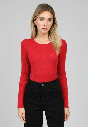 Femme aux cheveux blonds portant un haut rouge à manches longues côtelé ajusté et un pantalon noir taille haute avec des paillettes subtiles, debout devant un fond gris.