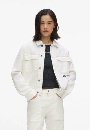 Kobieta w białej kurtce i spodniach dżinsowych z czarnym krótkim topem, stojąca z rękami w kieszeniach na jednolitym tle.