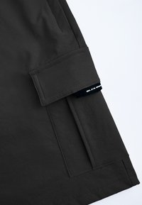 Donkergrijze stof met een gladde textuur en een opgerolde manchette. Bevat een zijzak en een label met de tekst "BLK AND WHITE" in het zwart.