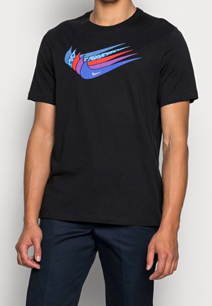 Mężczyzna ubrany w czarną koszulkę z krótkim rękawem z wielobarwnym wzorem Nike swoosh na piersi oraz ciemne spodnie.