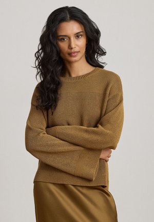 Frau mit langen lockigen Haaren, die einen braunen Strickpullover und einen passenden braunen Satinhrock trägt, steht mit verschränkten Armen vor einem neutralen Hintergrund.