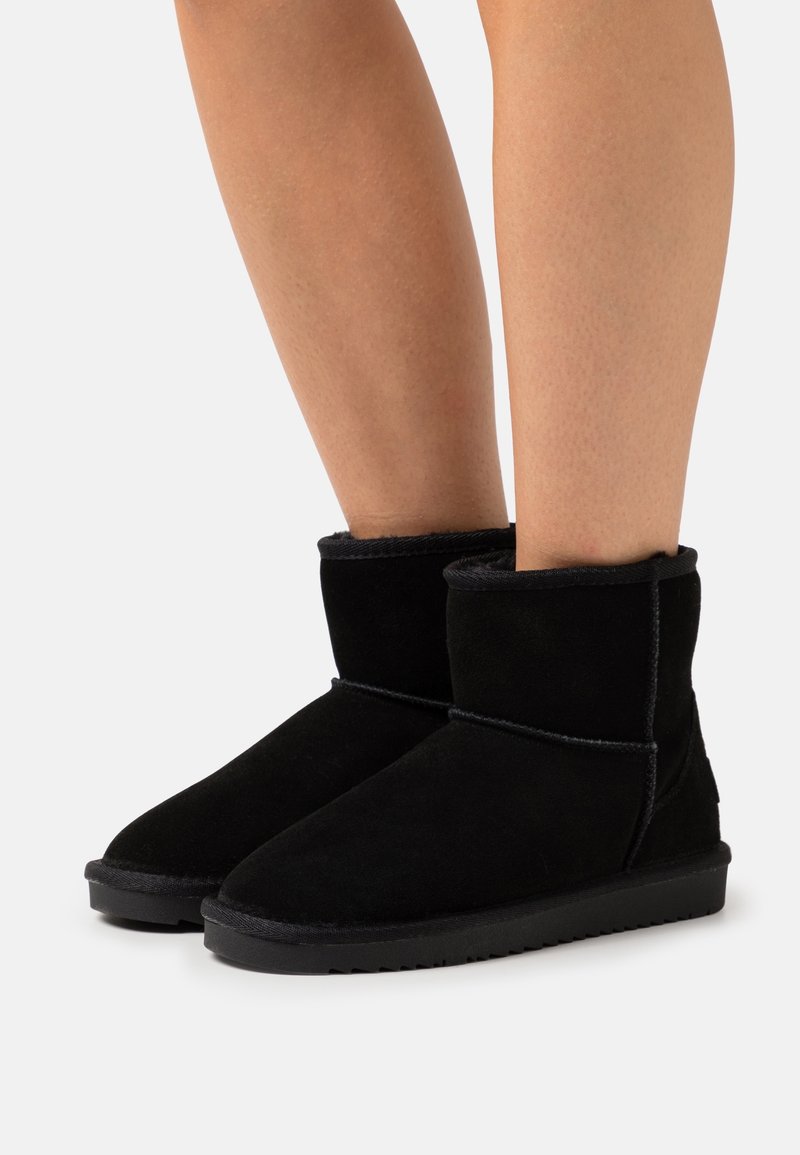 Bottes en daim noir montant jusqu'à la cheville avec semelles en caoutchouc plates portées sur des jambes nues sur un fond clair uni.