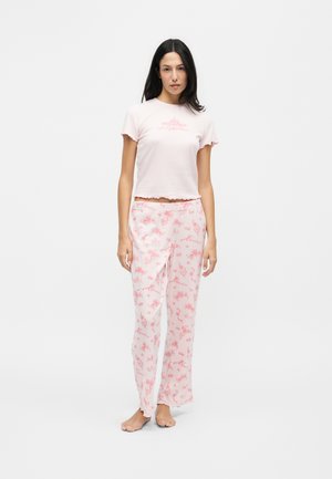 Femme debout pieds nus portant un haut rose clair à manches courtes et un pantalon ample rose à motifs floraux assortis sur fond blanc.