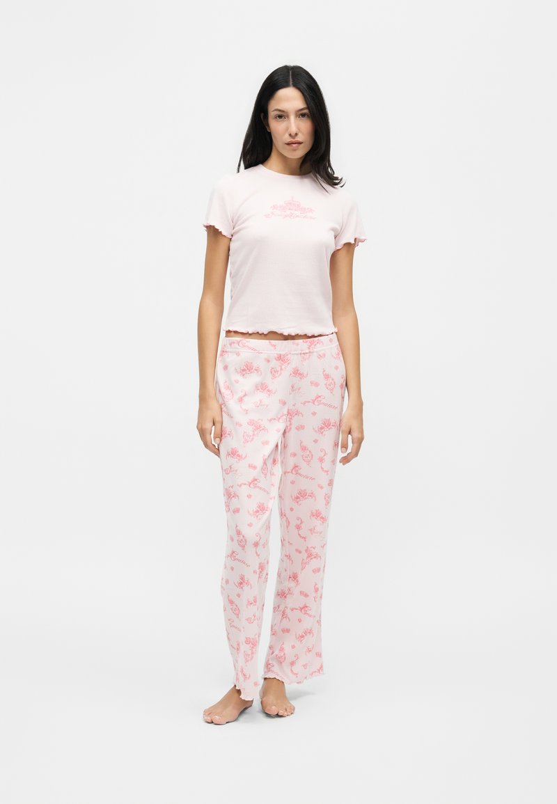 Femme debout pieds nus portant un haut rose clair à manches courtes et un pantalon ample rose à motifs floraux assortis sur fond blanc.