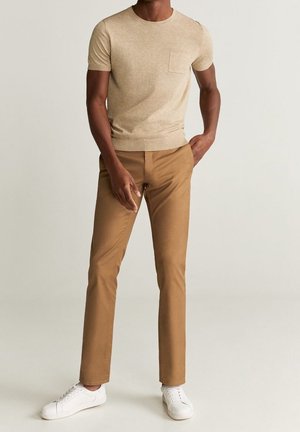 Homme portant une chemise beige à manches courtes, un pantalon marron clair et des baskets blanches, debout avec une main dans la poche.