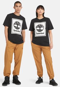 Zwarte katoenen t-shirts met een wit Timberland-logo, gecombineerd met tan kleurige elastische broeken; beide modellen dragen zwarte laarzen met een gestructureerd oppervlak.