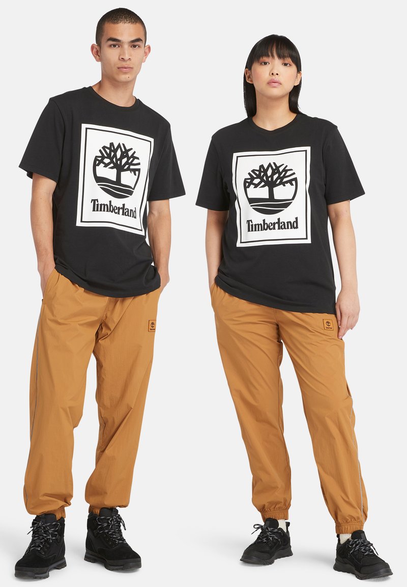 Zwarte katoenen t-shirts met een wit Timberland-logo, gecombineerd met tan kleurige elastische broeken; beide modellen dragen zwarte laarzen met een gestructureerd oppervlak.