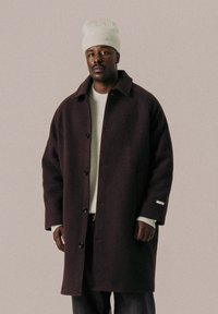 Maroon wool coat with large buttons, side pockets, and a relaxed fit. Paired with a white knit sweater and a light-colored beanie.