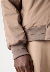 Main à peau foncée portant une veste beige à poignets et ceinture côtelés, reposant contre un pantalon beige assorti.