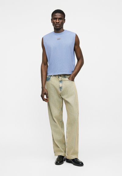 Νεαρός άνδρας που φοράει αμάνικη ανοιχτό μπλε μπλούζα, μπεζ τζιν φαρδιάς γραμμής και μαύρα παπούτσια, στέκεται με το ένα χέρι στην τσέπη μπροστά σε λευκό φόντο.