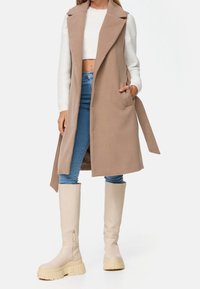 Longue veste beige avec col, devant ouvert et poches latérales, associée à un jean bleu clair et à des bottes à plateforme beige clair montant jusqu'aux genoux.