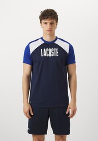 Mornarska atletika majica z belimi ramenskimi detajli, s poudarjeno "LACOSTE" blagovno znamko v beli barvi. V kompletu z usklajenimi mornarskimi kratkimi hlačami, obema iz gladkega materiala.