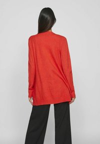 Lång röd tröja med lös passform, ribbad nederkant och långa ärmar. Bärs över svarta vida byxor som visar en jämn textur.