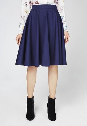 Žena stojící před jednobarevným pozadím, oblečená v tmavě modré sukně do kolen s rozšířeným střihem, květované halence s dlouhými rukávy a černých kotníkových botách.