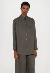 Camicia grigia con bottoni, maniche lunghe, taschino sul petto e un tessuto leggermente testurizzato. Presenta bottoni metallici e una vestibilità rilassata.