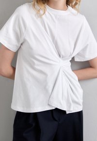 Bílý bavlněný tričko s uzlovým předním designem, krátkými rukávy a volným střihem. Hladký povrch s jemným splývavým efektem.